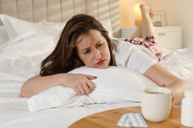 Photo of Unhappy woman with cup of coffee and pills suffering from hangover on bed at home