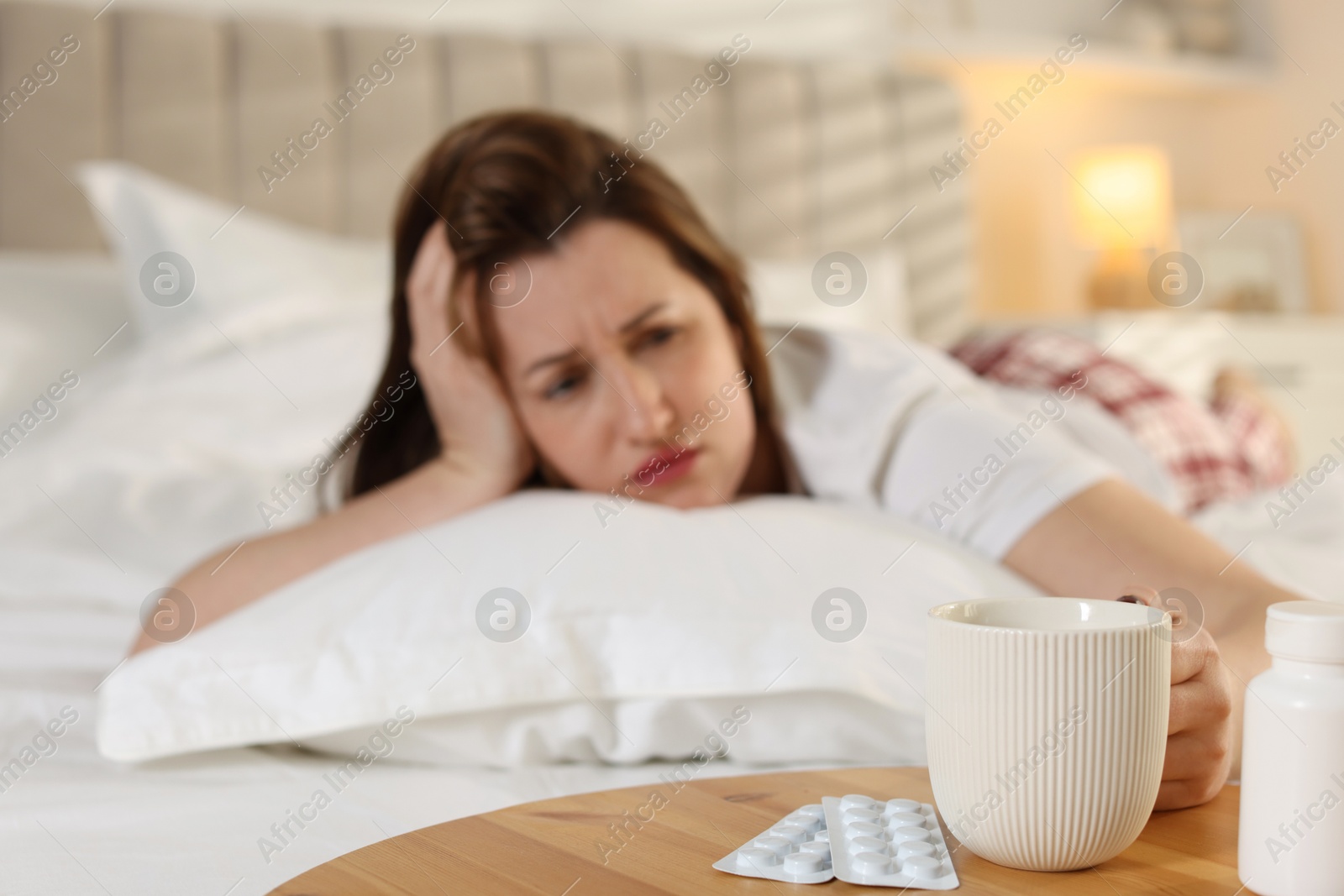 Photo of Unhappy woman with cup of coffee and pills suffering from hangover on bed at home, selective focus