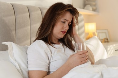 Photo of Unhappy woman with glass of water suffering from hangover on bed at home