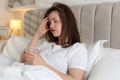 Unhappy woman with glass of water suffering from hangover on bed at home