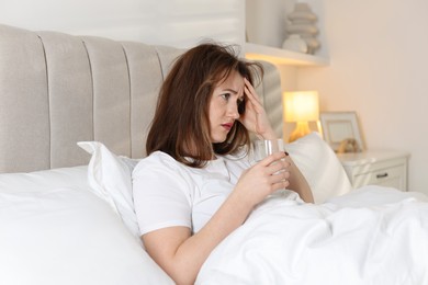 Unhappy woman with glass of water suffering from hangover on bed at home