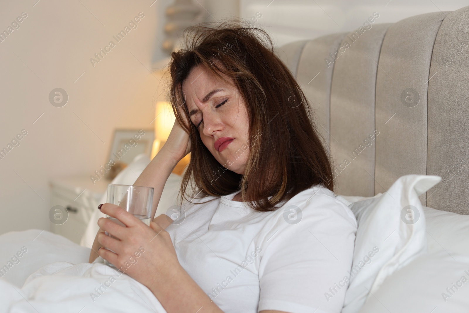 Photo of Unhappy woman with glass of water suffering from hangover on bed at home