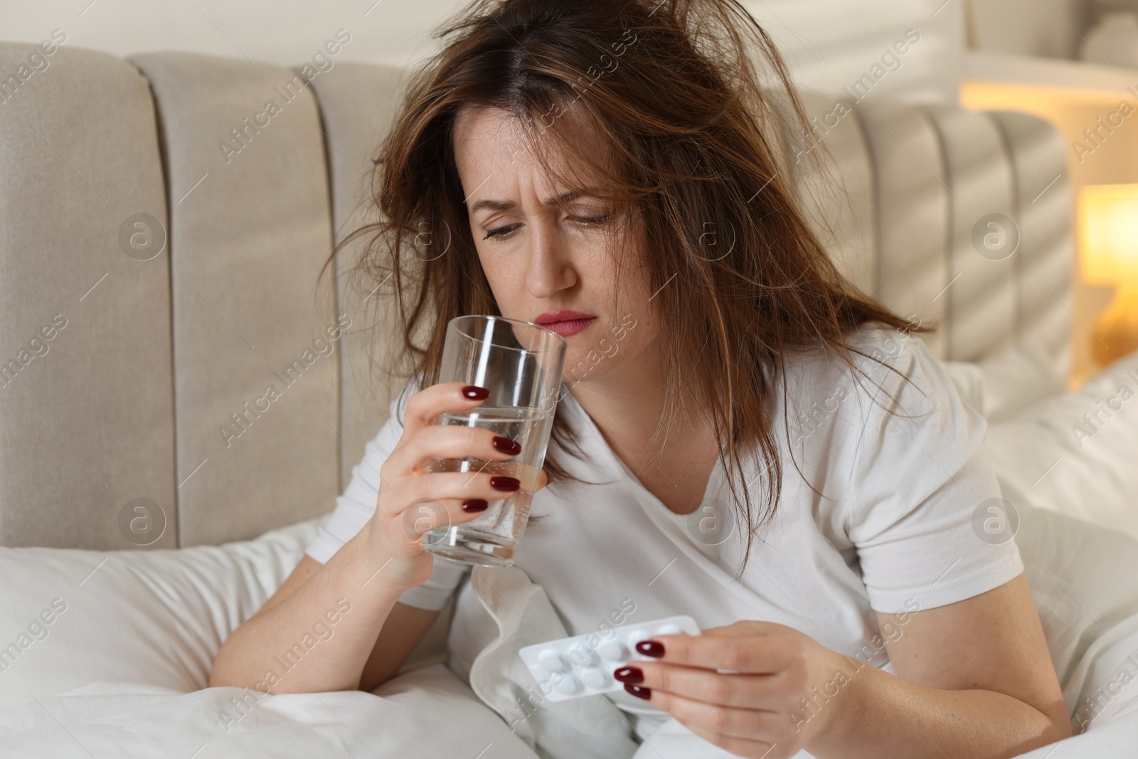 Photo of Unhappy woman with pills and glass of water suffering from hangover on bed at home