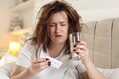 Unhappy woman with pills and glass of water suffering from hangover on bed at home