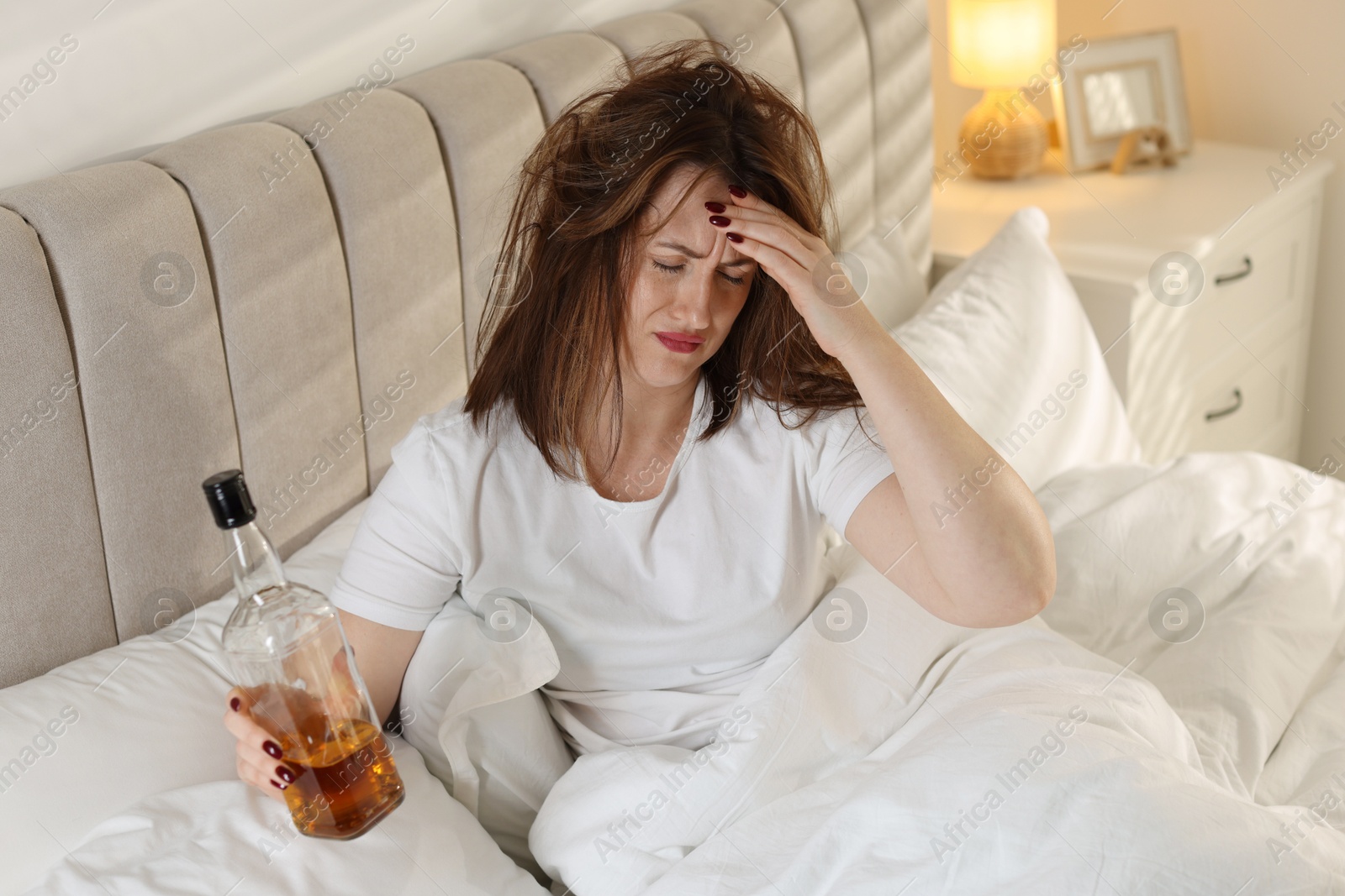 Photo of Unhappy woman with bottle of whiskey suffering from hangover on bed at home