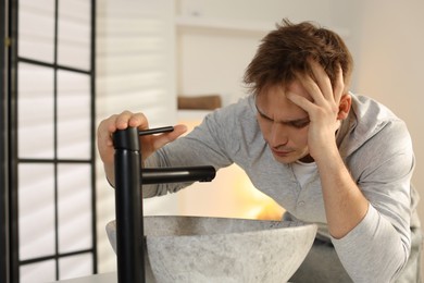 Photo of Unhappy young man suffering from hangover near sink in bathroom