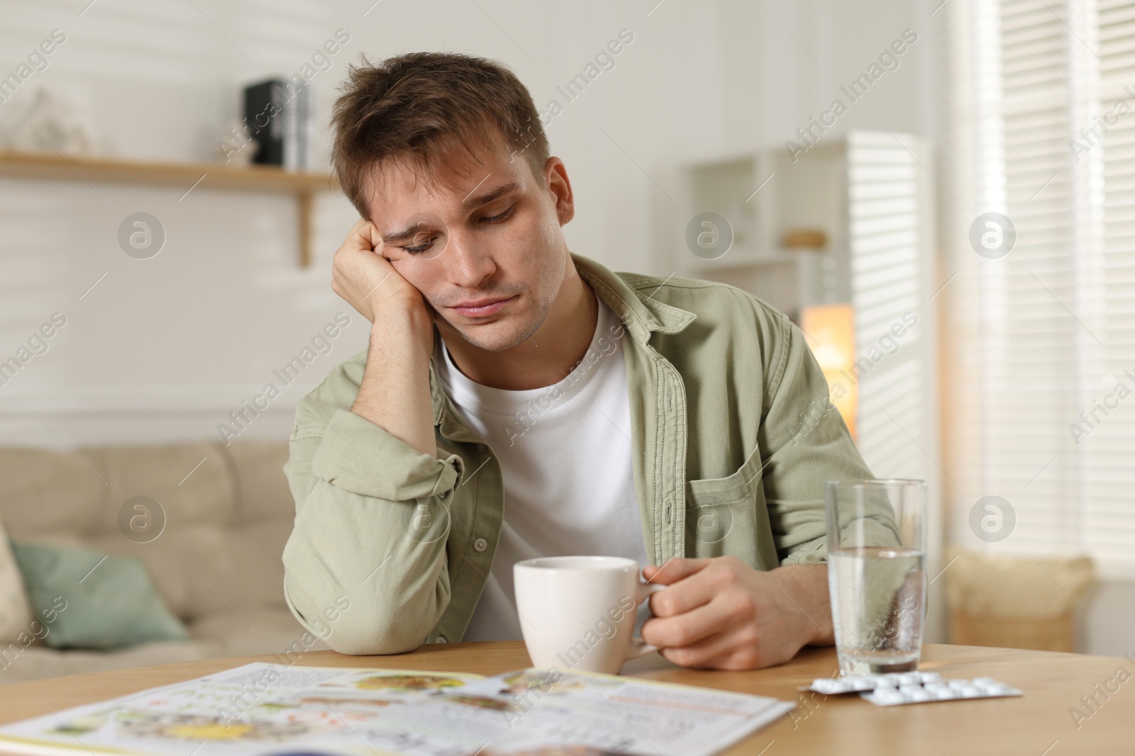 Photo of Suffering from hangover. Unhappy young man with cup of coffee sitting at wooden table indoors