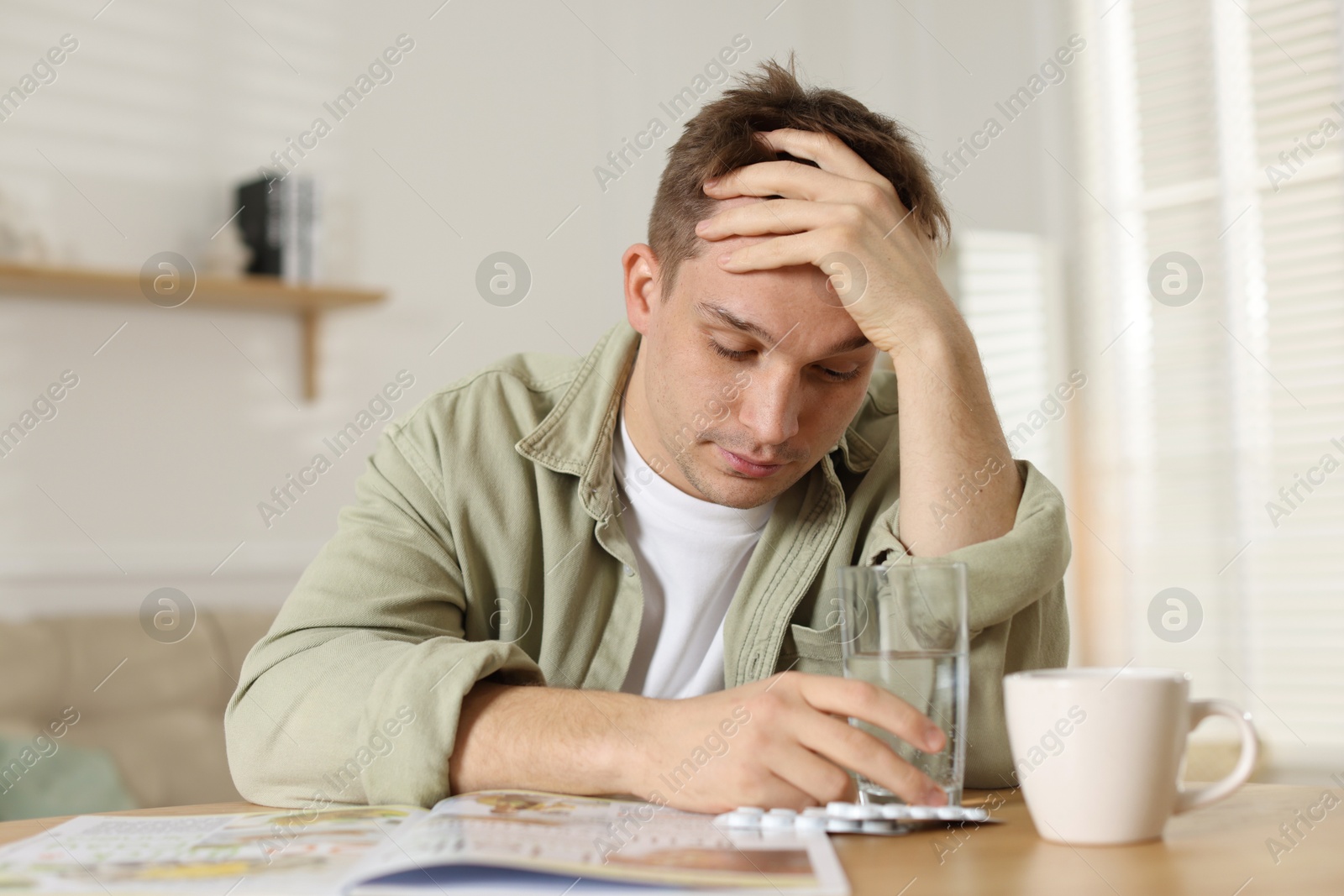 Photo of Suffering from hangover. Unhappy young man sitting at wooden table indoors