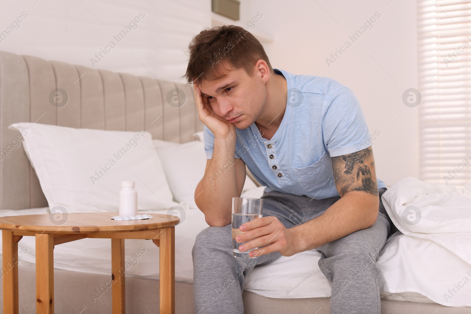 Photo of Unhappy young man with glass of water suffering from hangover on bed at home