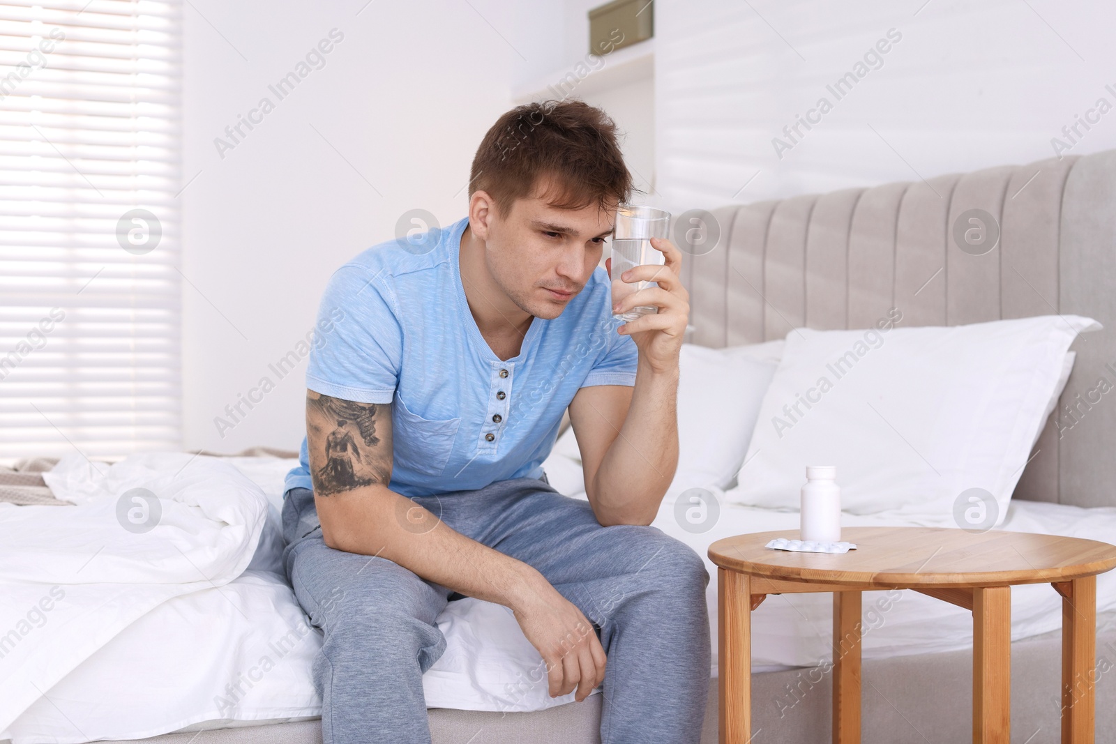 Photo of Unhappy young man with glass of water suffering from hangover on bed at home