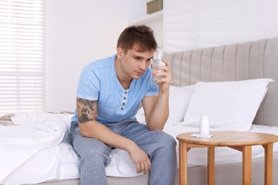 Unhappy young man with glass of water suffering from hangover on bed at home