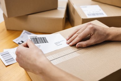 Photo of Man sticking shipping label with barcode on parcel at wooden table indoors, closeup