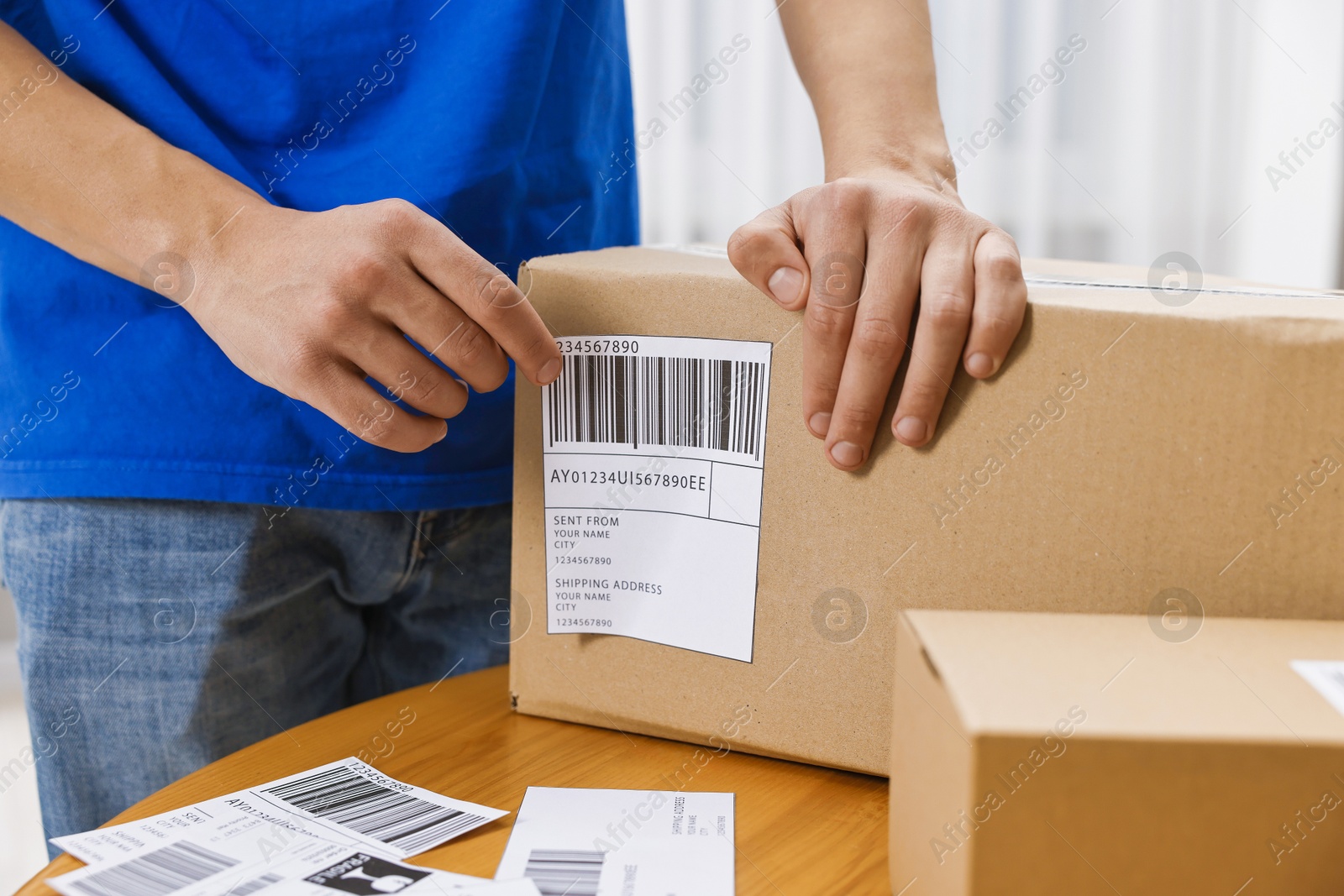 Photo of Man sticking shipping label with barcode on parcel at wooden table indoors, closeup