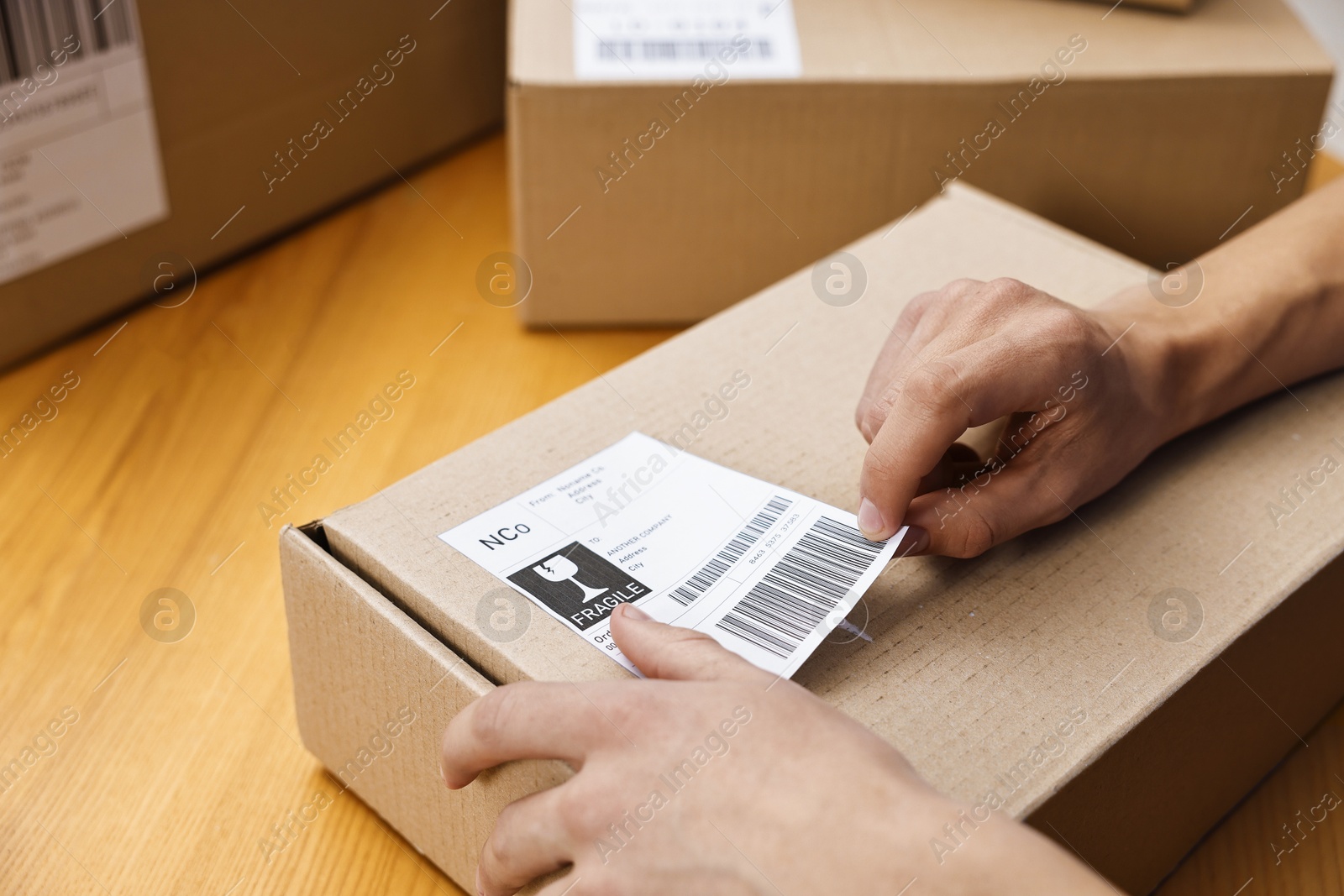 Photo of Man sticking shipping label with barcode on parcel at wooden table indoors, closeup