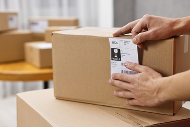 Photo of Man sticking shipping label with barcode on parcel indoors, closeup