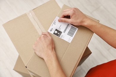 Photo of Man sticking shipping label with barcode on parcel indoors, above view