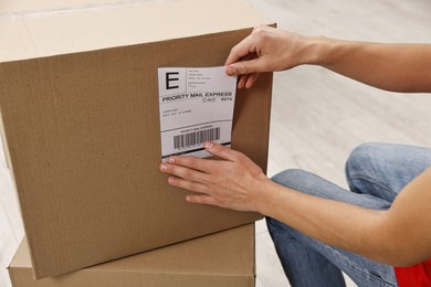 Photo of Man sticking shipping label with barcode on parcel indoors, closeup