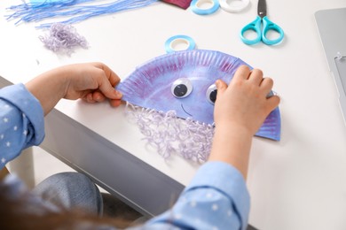 Photo of Little girl making paper jellyfish at white table, closeup. Child handmade craft