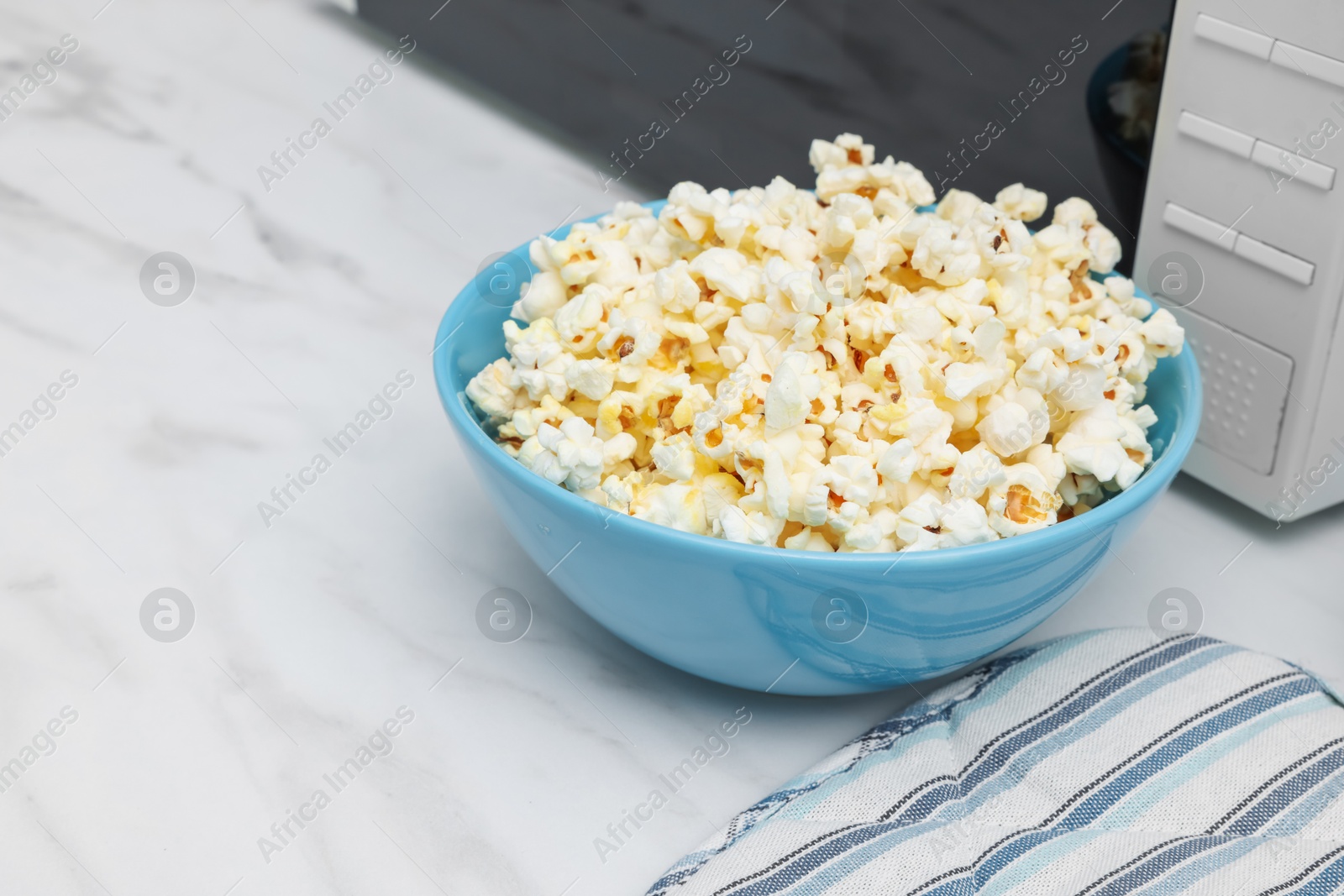 Photo of Tasty popcorn in bowl near microwave oven on white marble table, closeup. Space for text