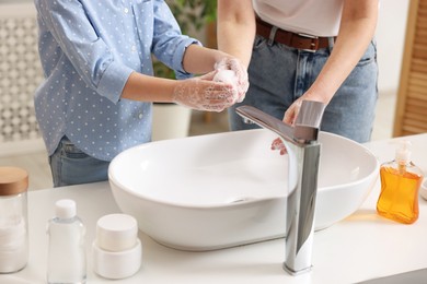 Mother and daughter washing their hands above sink indoors, closeup