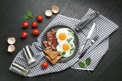 Photo of Tasty fried eggs, bacon and pieces of toast served on black table, flat lay