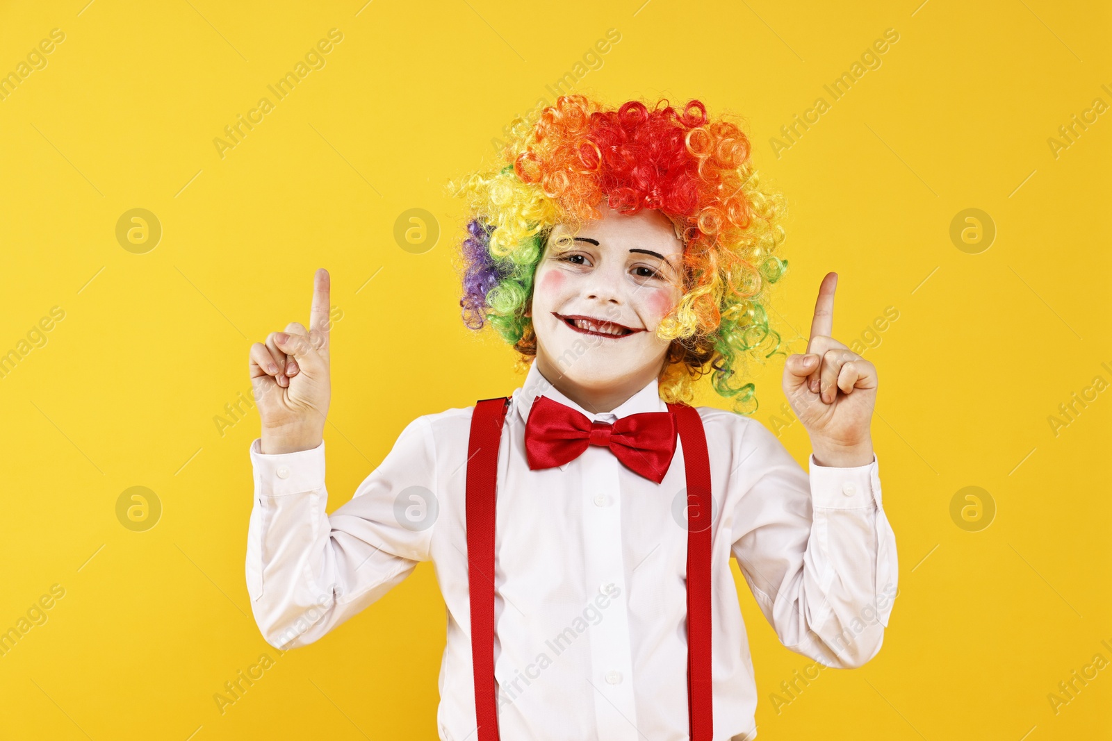 Photo of Happy little boy dressed like clown pointing upwards on yellow background. Surprise party