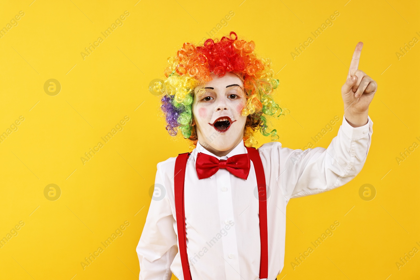 Photo of Emotional little boy dressed like clown pointing upwards on yellow background, space for text. Surprise party