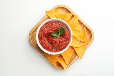 Delicious spicy salsa sauce in bowl and nacho chips on white wooden table, top view