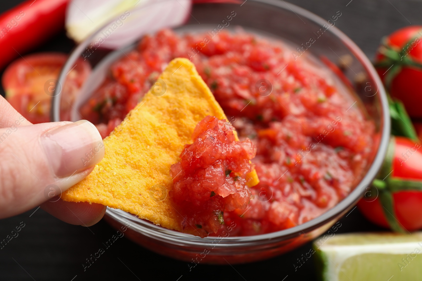 Photo of Woman dipping nacho chip into spicy salsa sauce at table, closeup