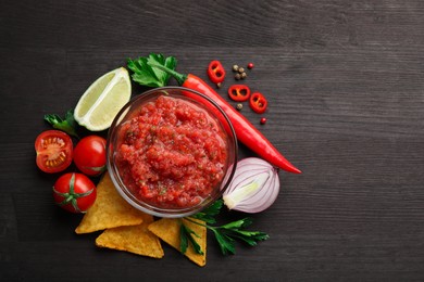 Spicy salsa sauce in bowl, nachos and ingredients on wooden table, flat lay. Space for text