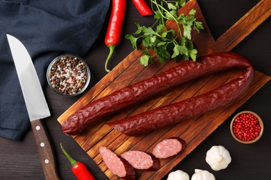 Photo of Delicious thin smoked sausage, spices and knife on dark wooden table, flat lay