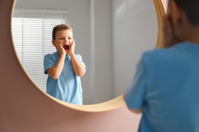 Photo of Cute little boy with missing tooth near mirror indoors