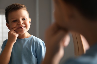 Photo of Cute little boy pointing at his missing tooth near mirror indoors