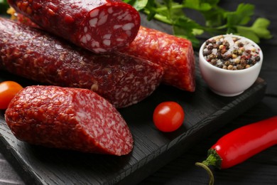 Photo of Different smoked sausages, tomato, chili pepper and peppercorns on black wooden table, closeup