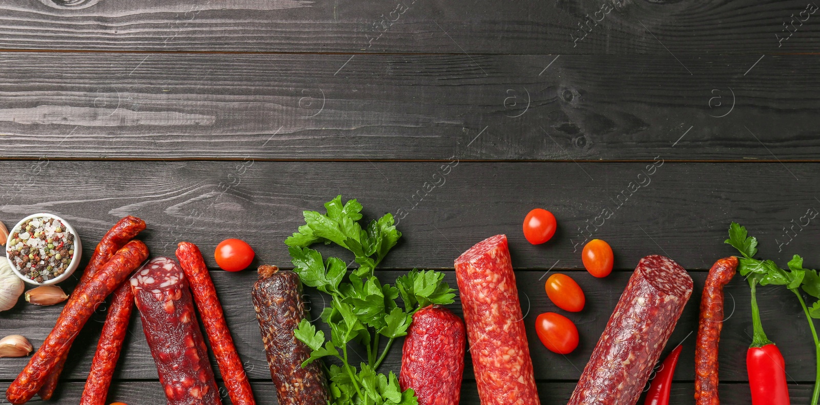 Photo of Different smoked sausages, tomatoes, parsley and chili pepper on black wooden table, flat lay. Space for text