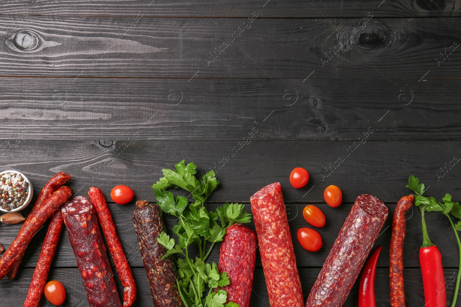 Photo of Different smoked sausages, tomatoes, parsley and chili pepper on black wooden table, flat lay. Space for text