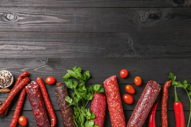 Photo of Different smoked sausages, tomatoes, parsley and chili pepper on black wooden table, flat lay. Space for text