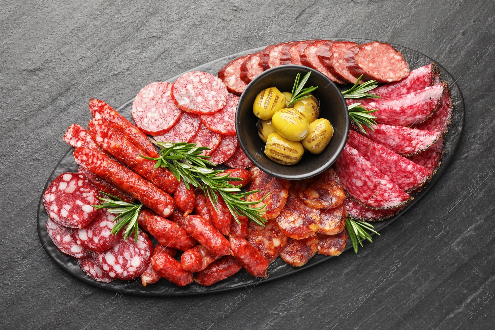 Photo of Different smoked sausages, olives and rosemary on black table, top view