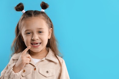 Photo of Cute little girl pointing at her missing tooth on light blue background, space for text. Waiting for tooth fairy