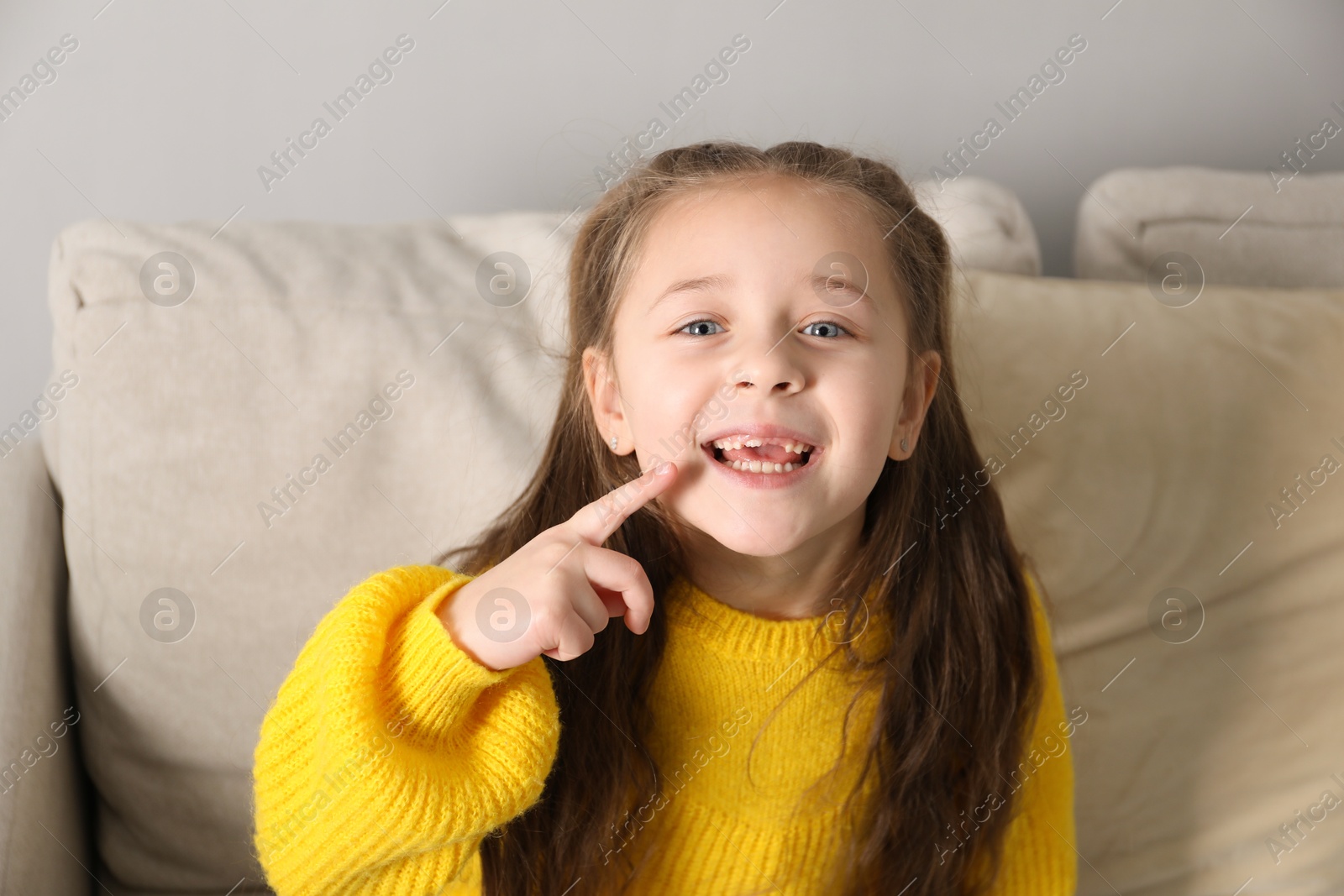 Photo of Cute little girl pointing at her missing tooth on sofa indoors. Waiting for tooth fairy