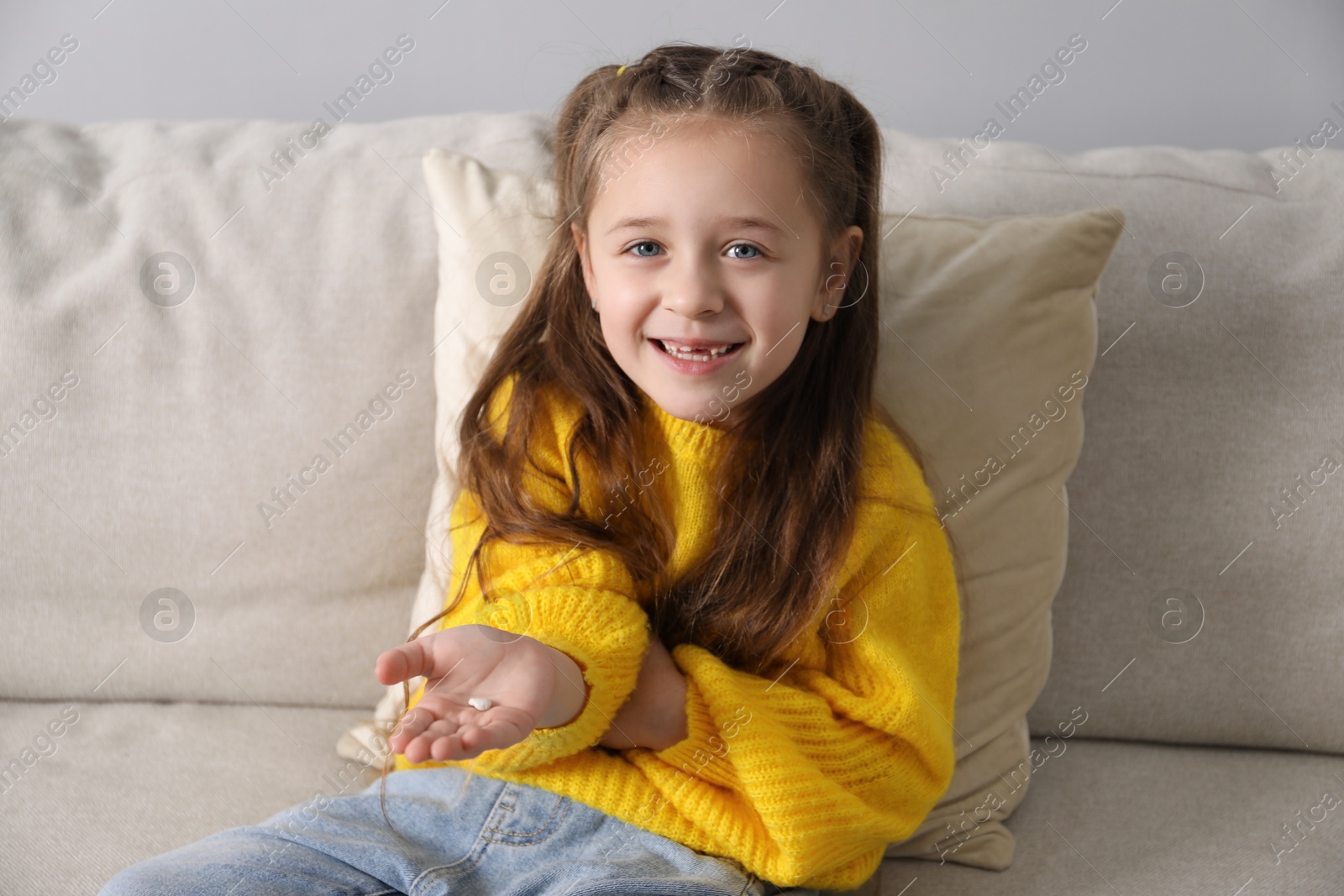 Photo of Cute little girl with missing tooth on sofa indoors. Waiting for tooth fairy