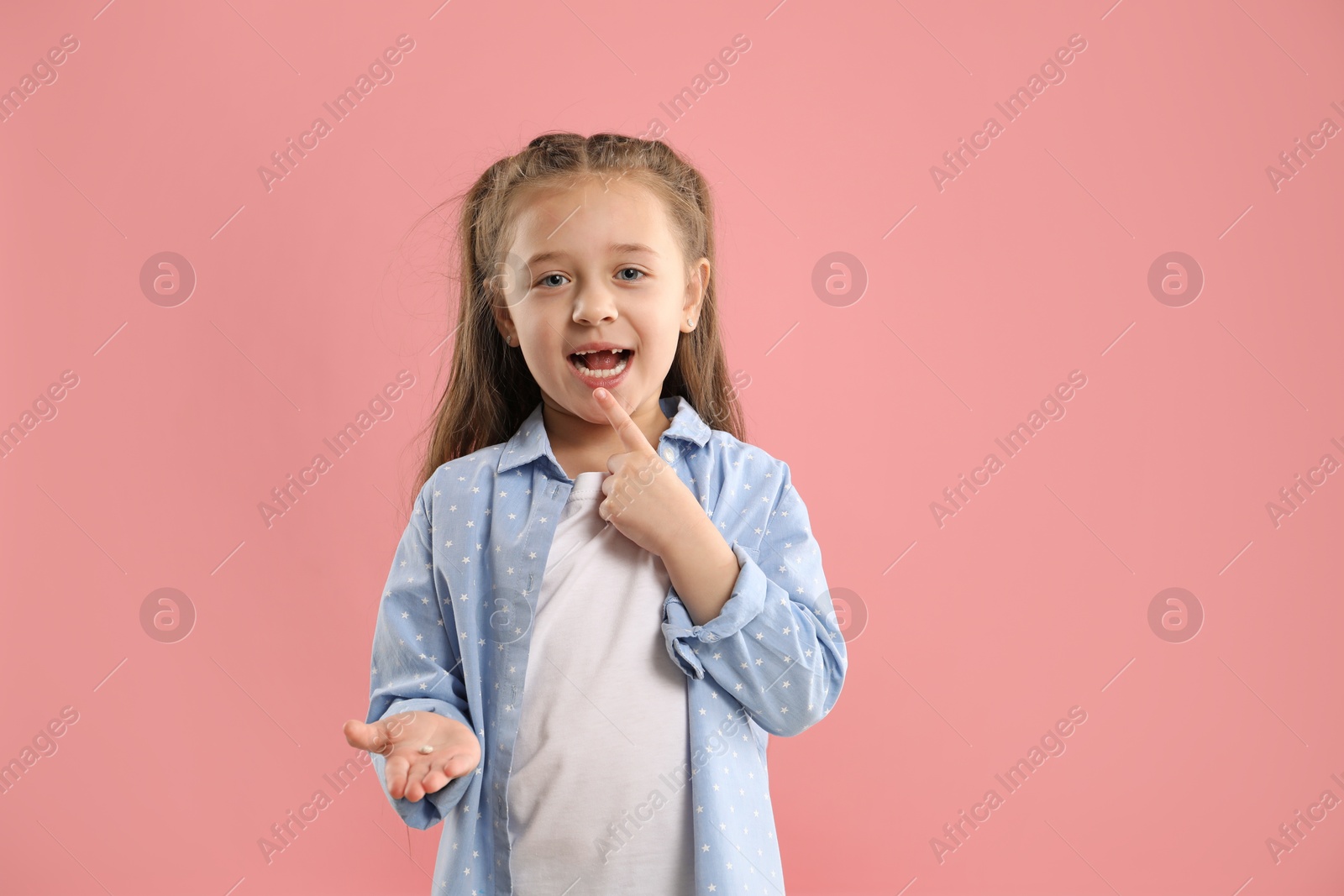 Photo of Cute little girl pointing at her missing tooth on pink background. Waiting for tooth fairy