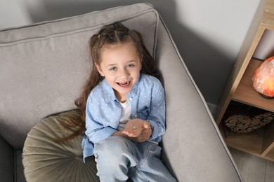 Photo of Cute little girl with missing tooth in armchair at home, above view. Waiting for tooth fairy