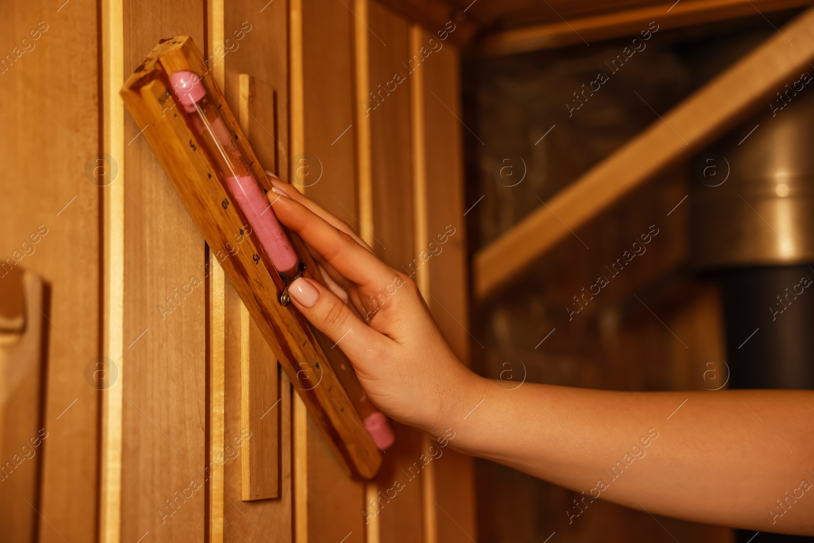 Photo of Woman turning hourglass on wooden wall in sauna, closeup