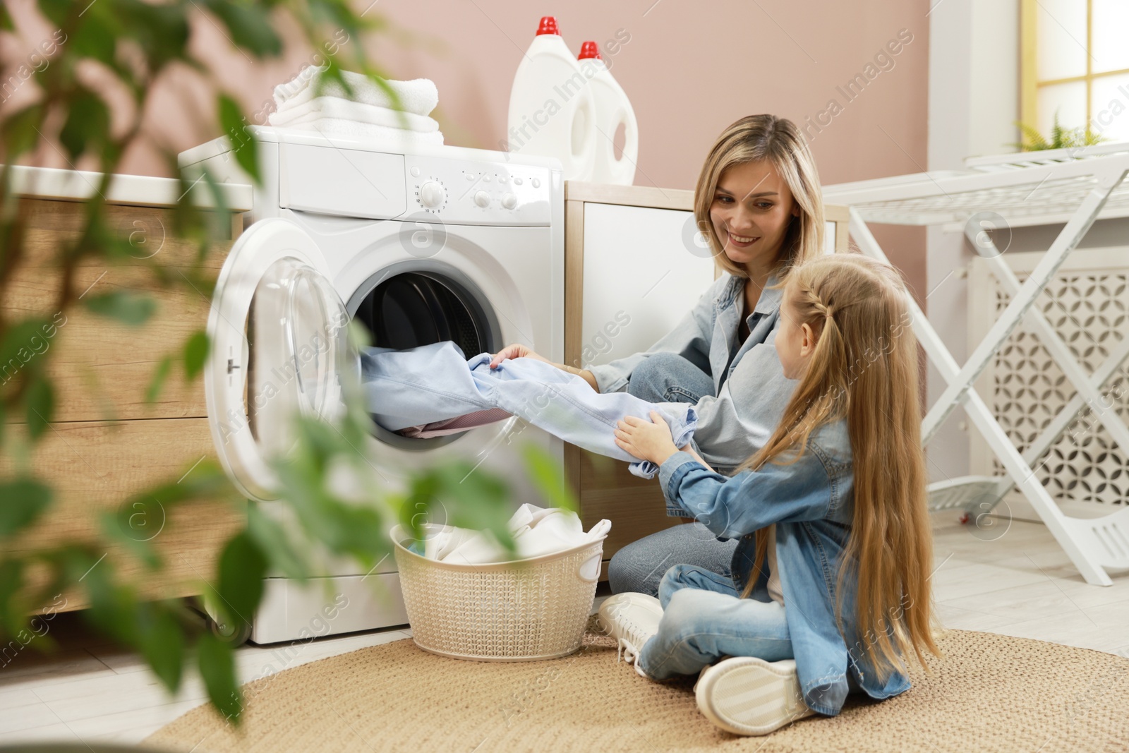 Photo of Mother and daughter with laundry basket loading washing machine together at home