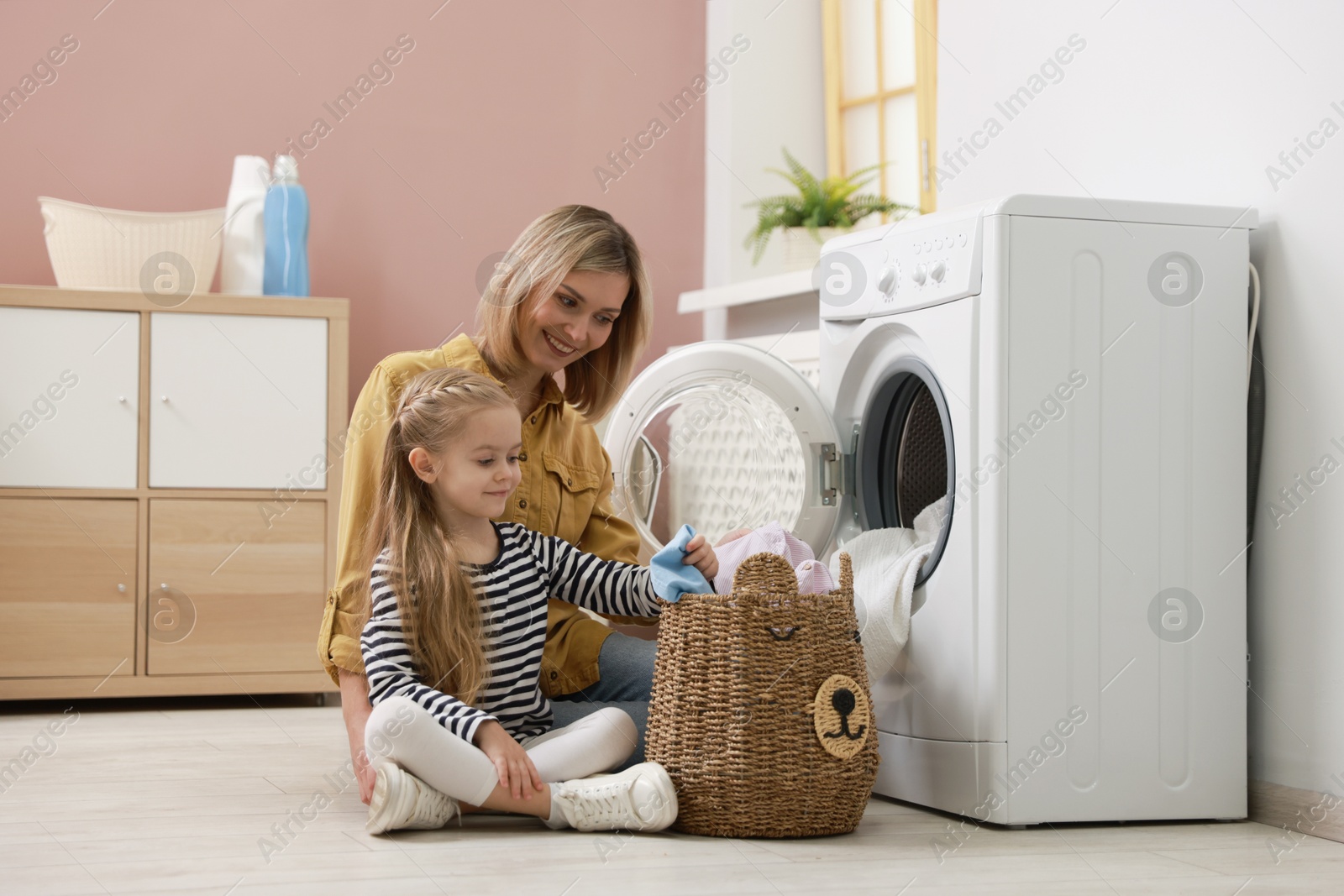 Photo of Mother and daughter with laundry basket loading washing machine together at home