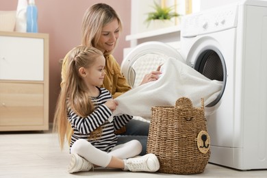 Photo of Mother and daughter with laundry basket loading washing machine together at home