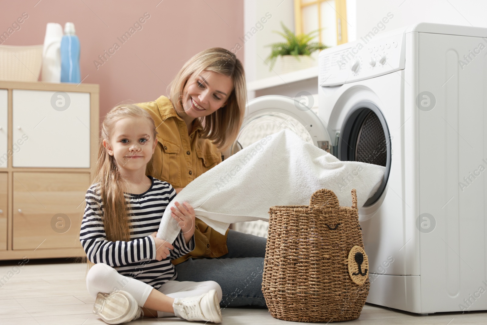 Photo of Mother and daughter with laundry basket loading washing machine together at home