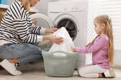 Mother and daughter with laundry basket loading washing machine together at home