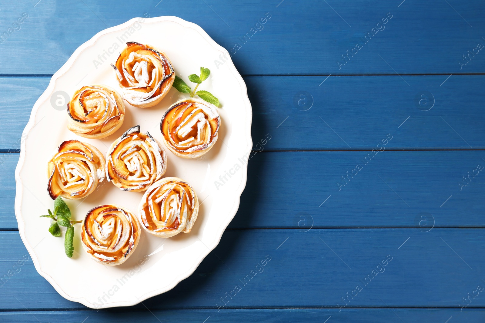 Photo of Tasty apple roses with powdered sugar served on blue wooden table, top view